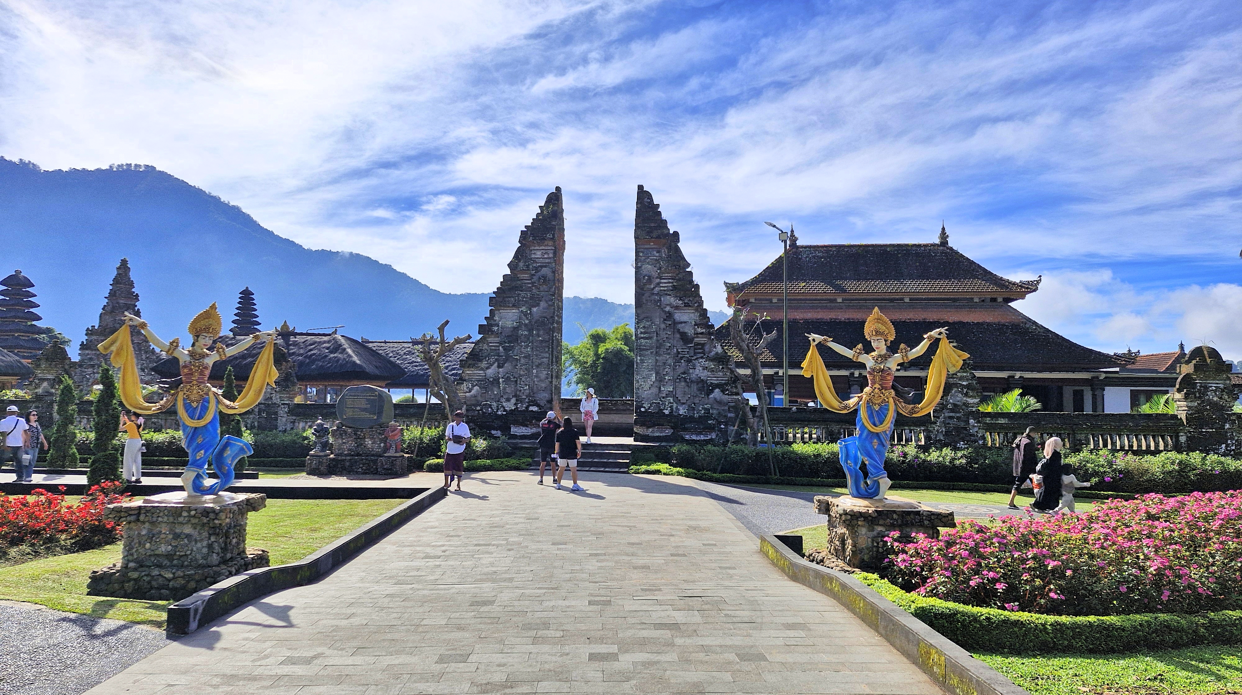 View of Ulun Danu Beratan Temple entrance with statues and floral decorations, set against a backdrop of mountains and blue skies.