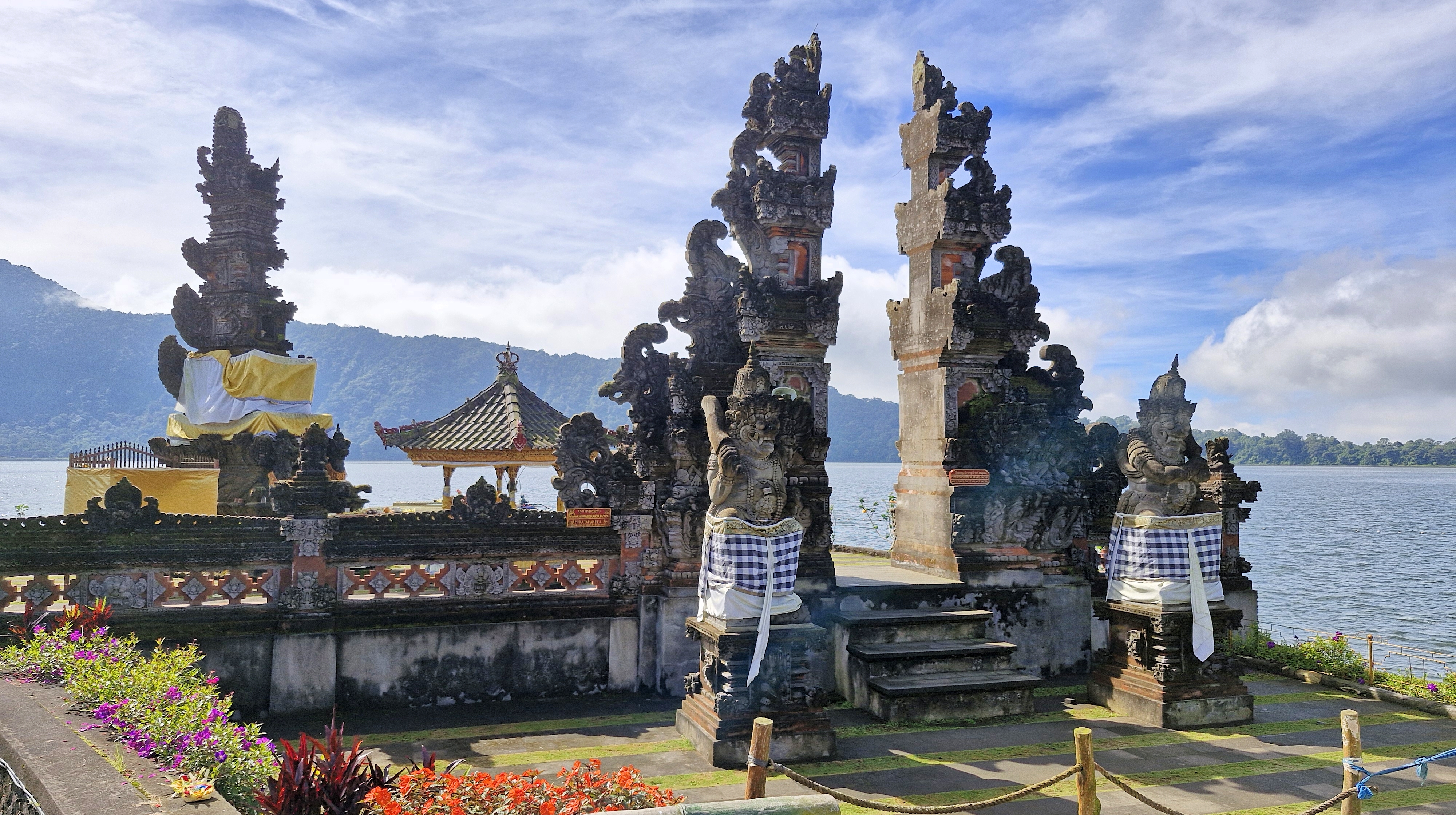 View of the Ulun Danu Beratan Temple, featuring intricate stone carvings and statues, located on the shores of Lake Beratan in Bali, against a backdrop of mountains and a blue sky.