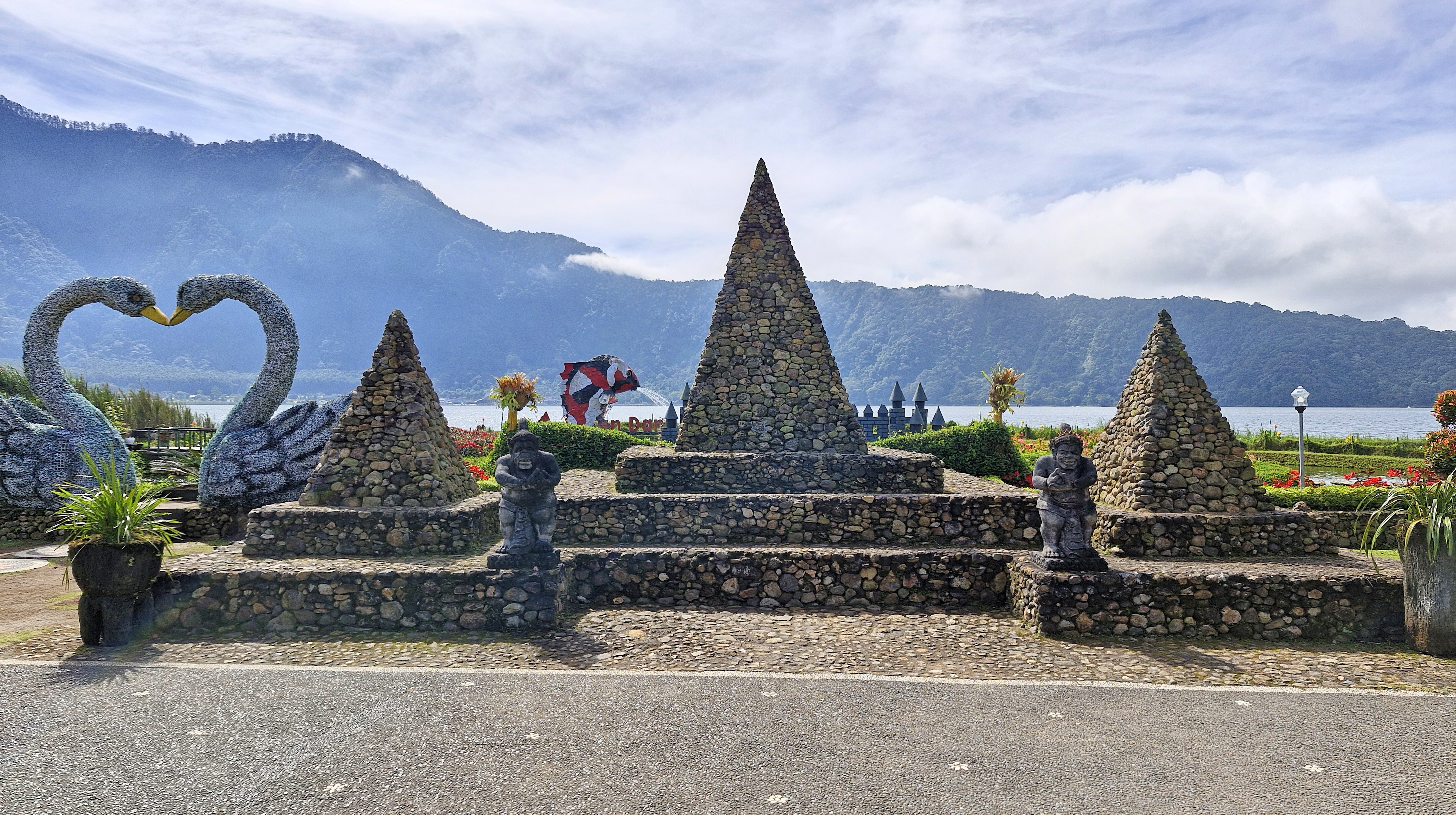 View of stone pyramids and decorative swan sculptures by a lake and mountains in the background.