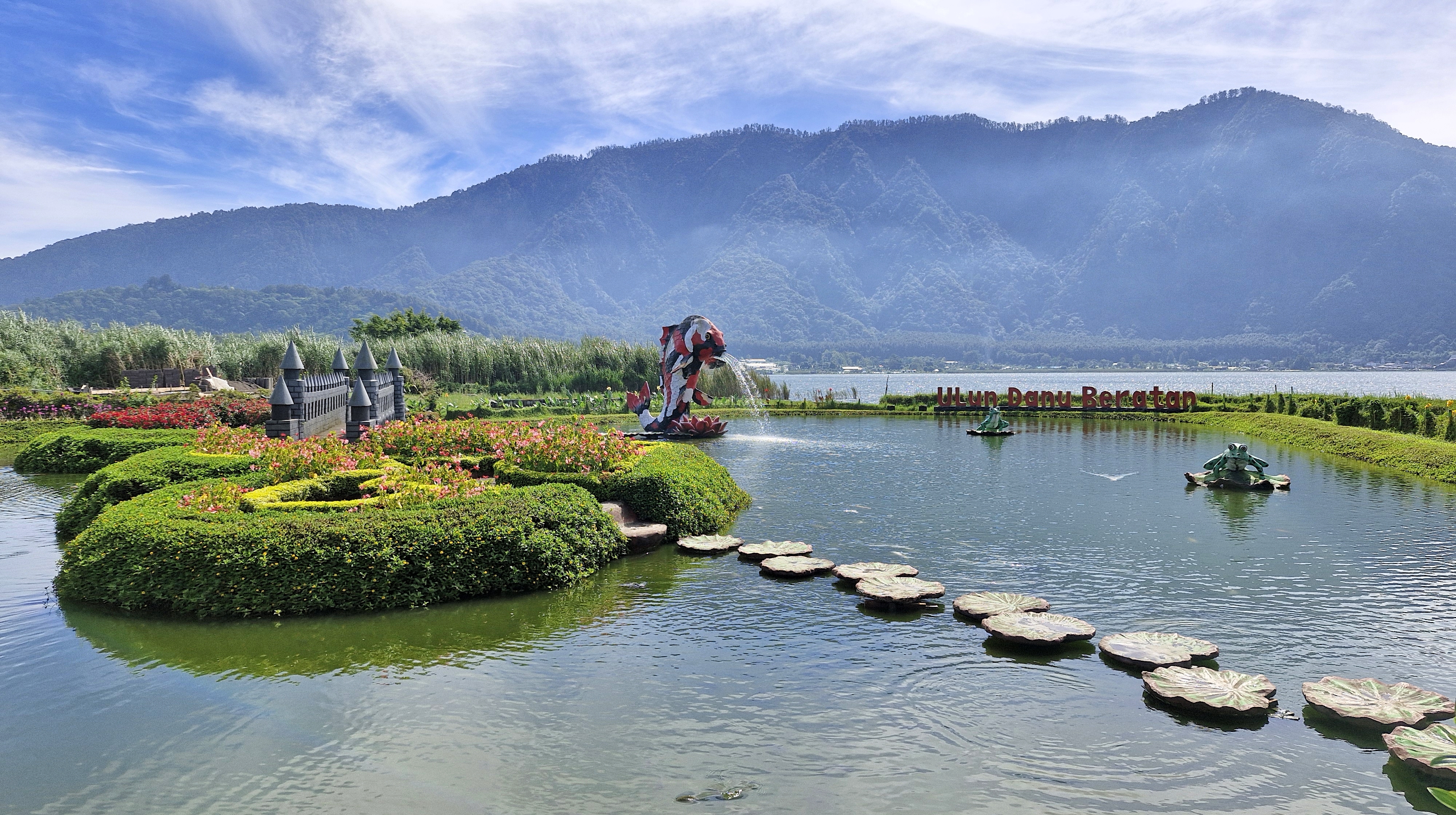 A scenic view of the Ulun Danu Beratan temple located on Lake Beratan, surrounded by lush greenery and mountains under a partly cloudy sky.
