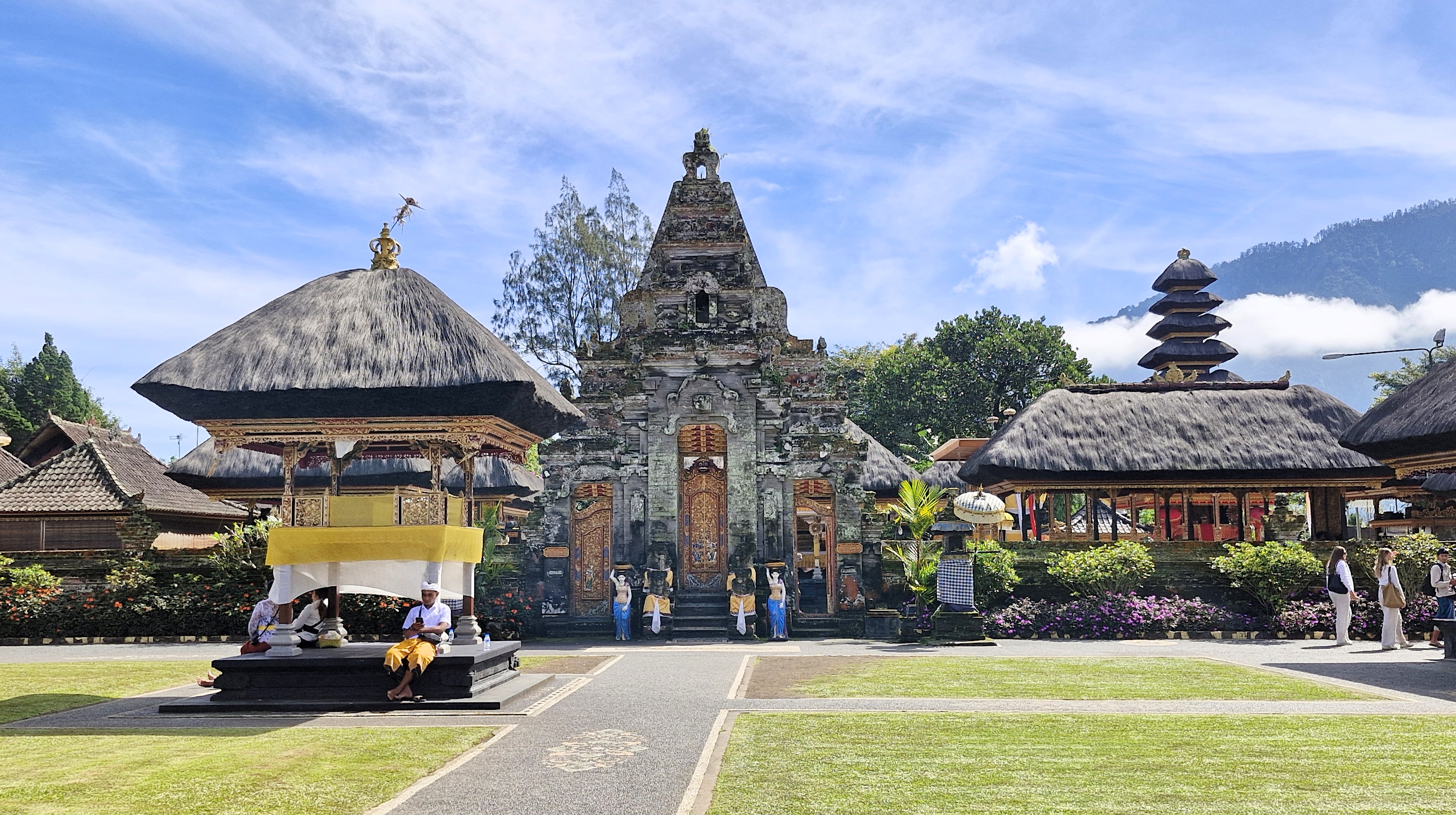 A panoramic view of the Ulun Danu Beratan temple in Bali, showcasing traditional Balinese architecture, lush gardens, and visitors exploring the sacred site under a clear blue sky.