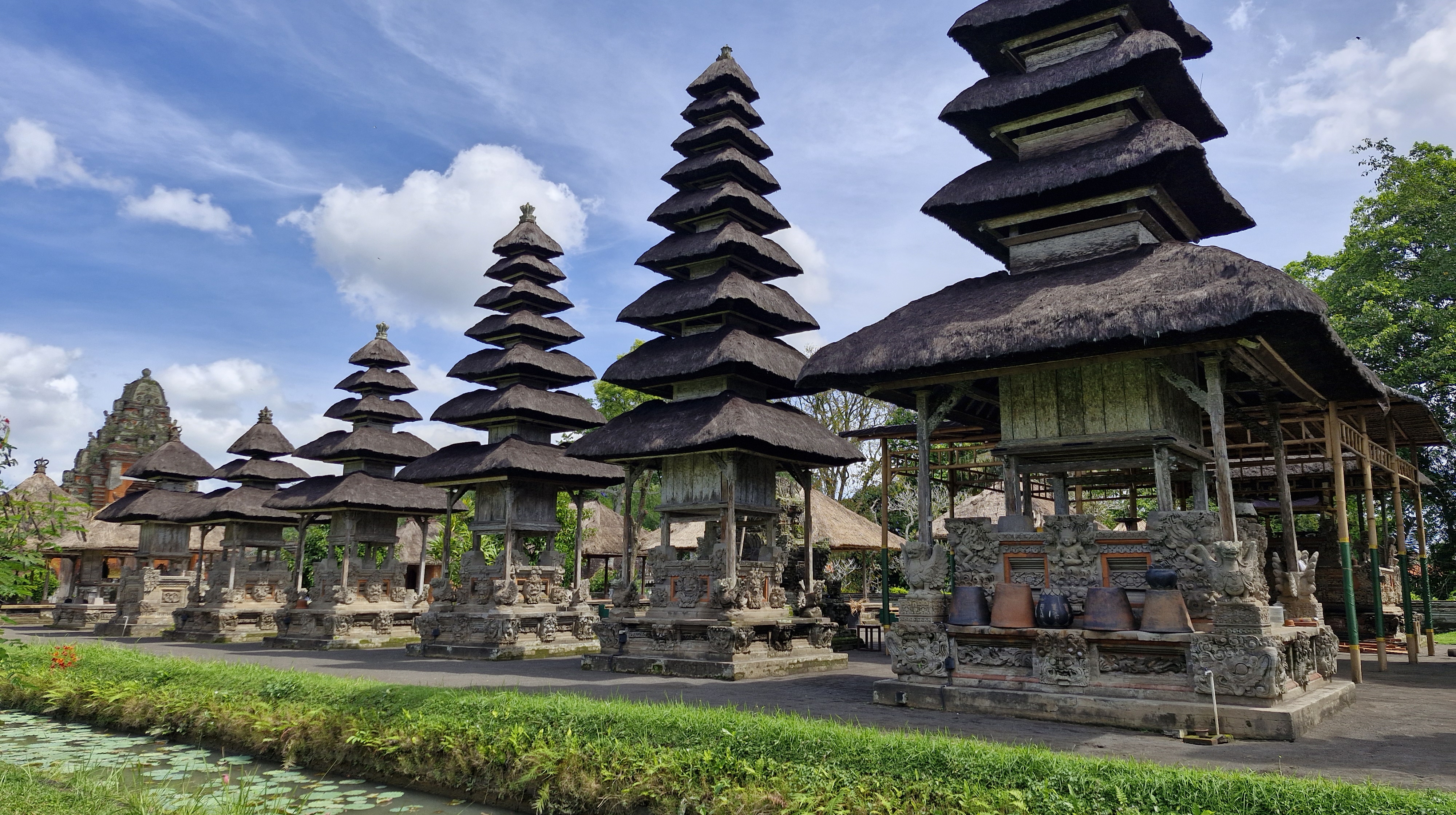 A view of the traditional Balinese temple structures with tiered roofs set against a blue sky, surrounded by lush greenery and a decorative stone wall.