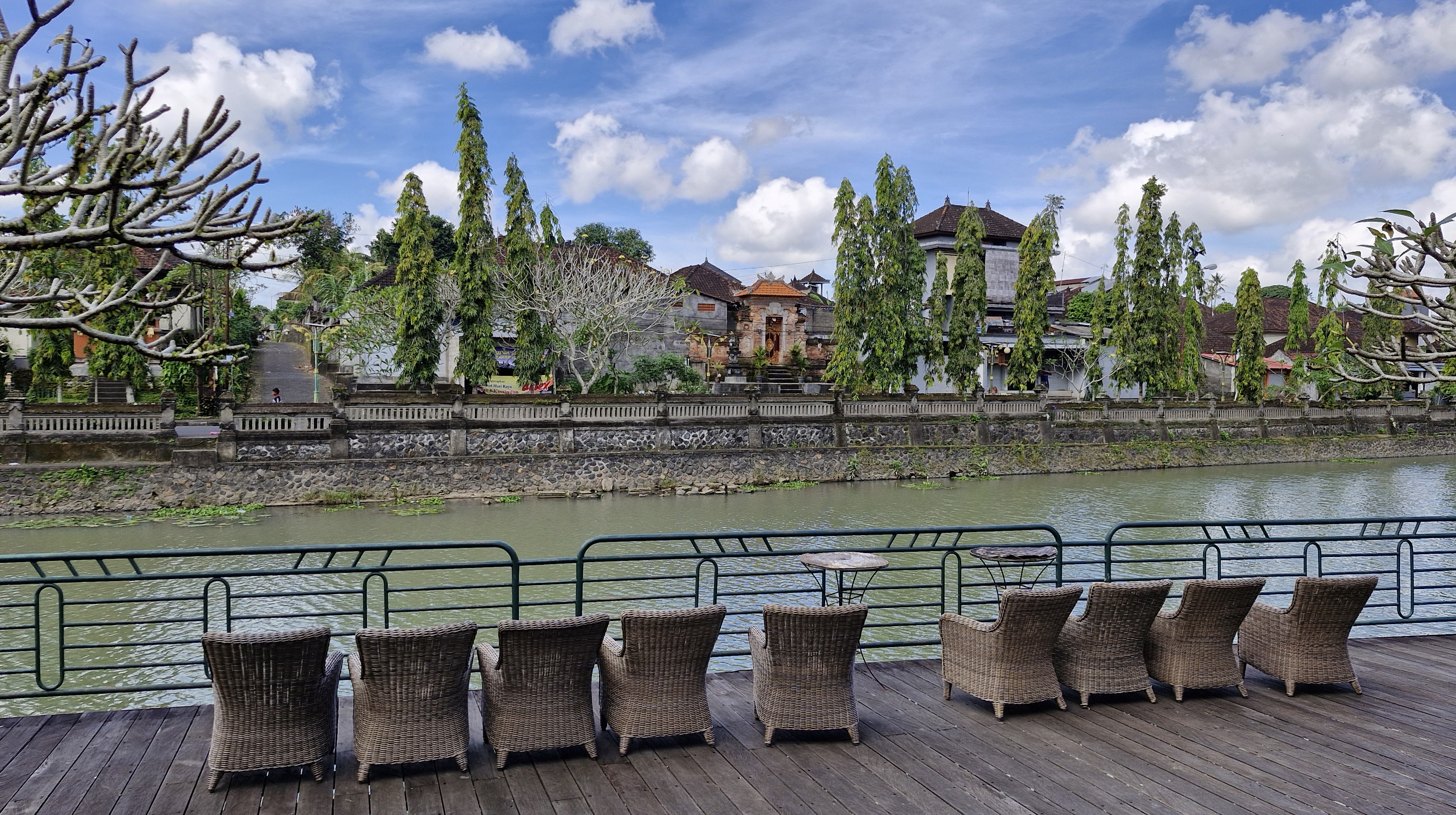 View of the river bordered by chairs and tables, with lush greenery and traditional buildings visible in the background under a blue sky.