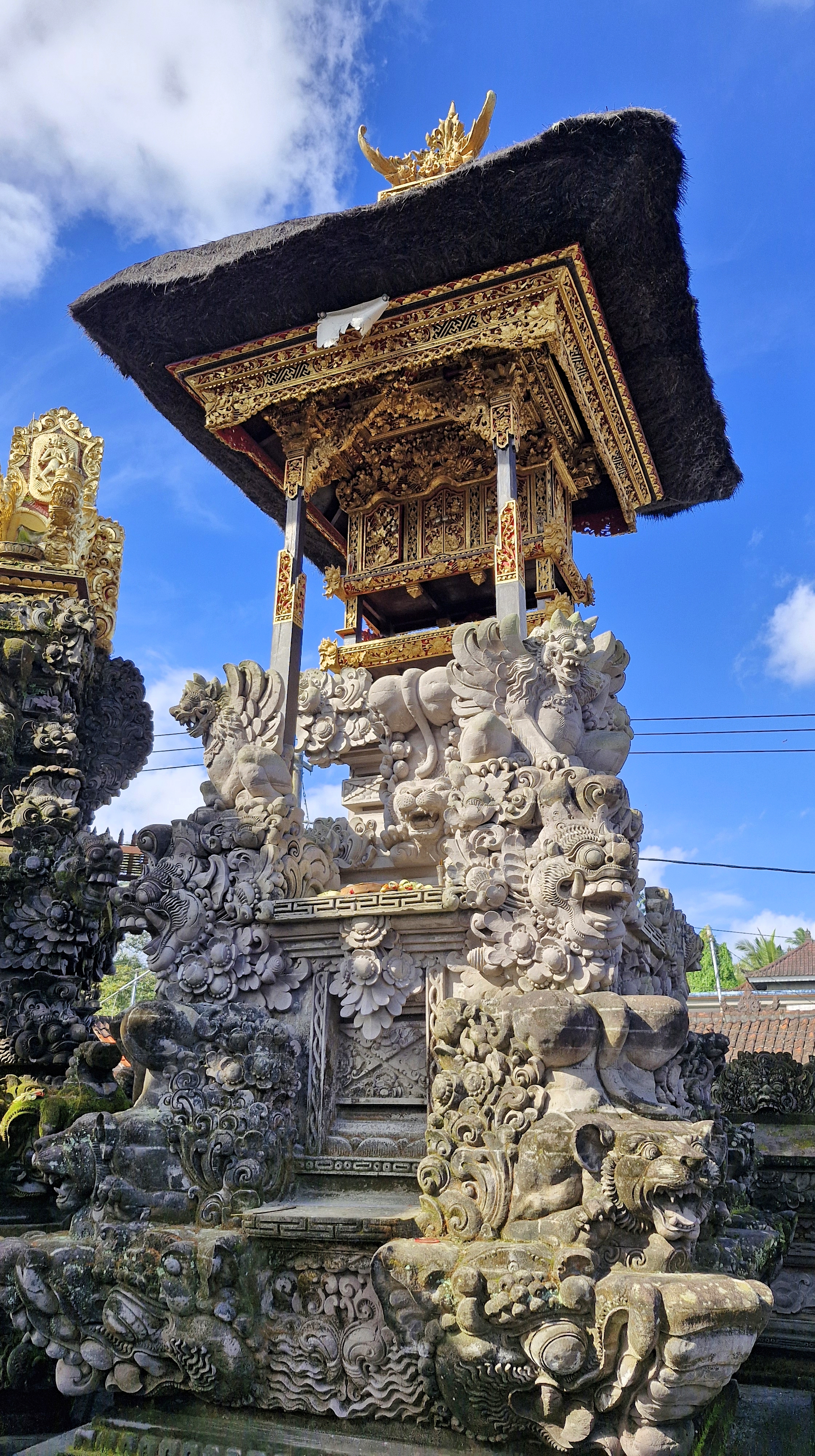 A detailed view of a traditional Balinese temple structure, featuring intricate carvings and a thatched roof under a blue sky.