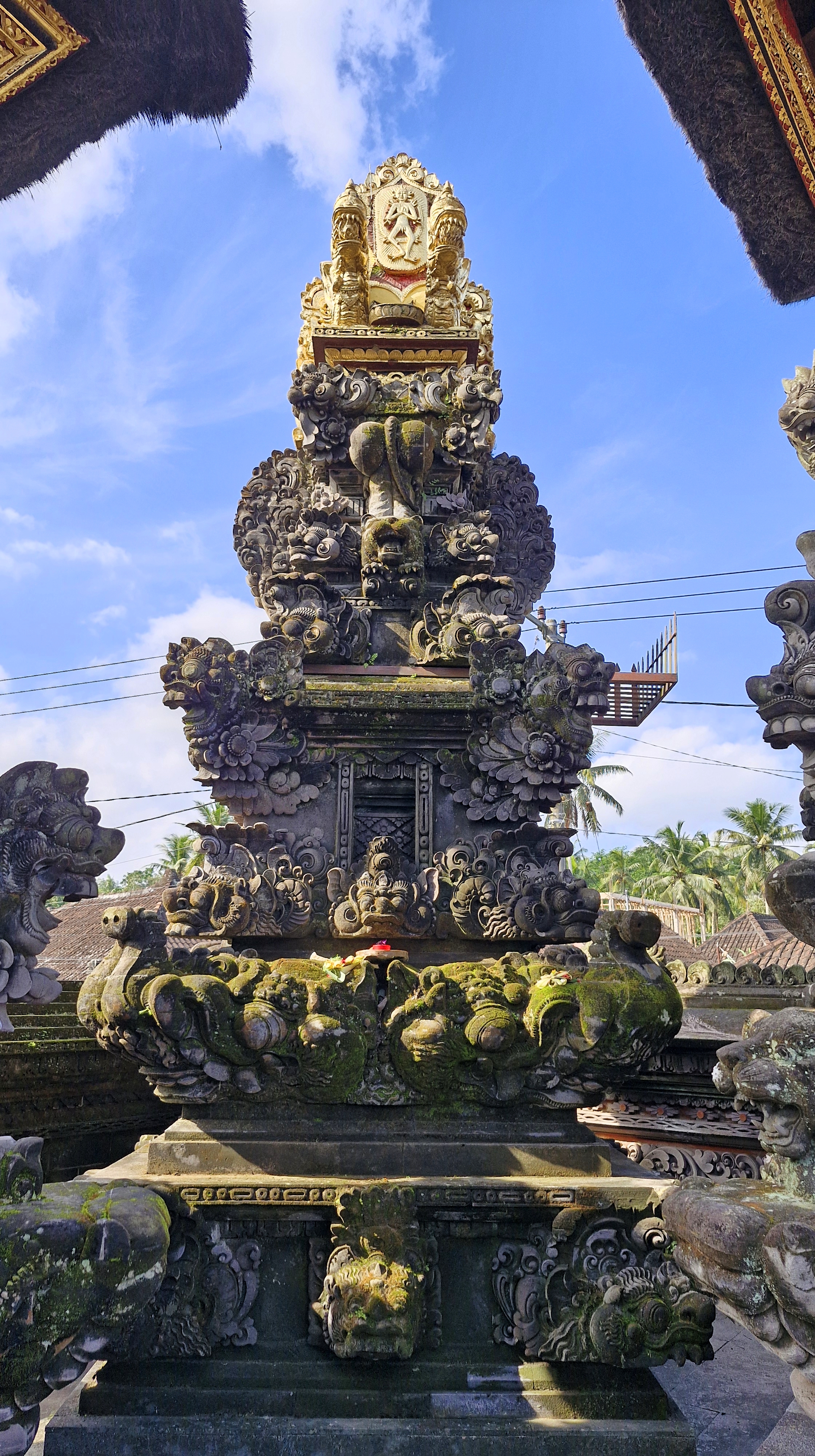 A decorative Balinese temple structure with intricate carvings and a statue at the top, set against a blue sky with fluffy clouds.