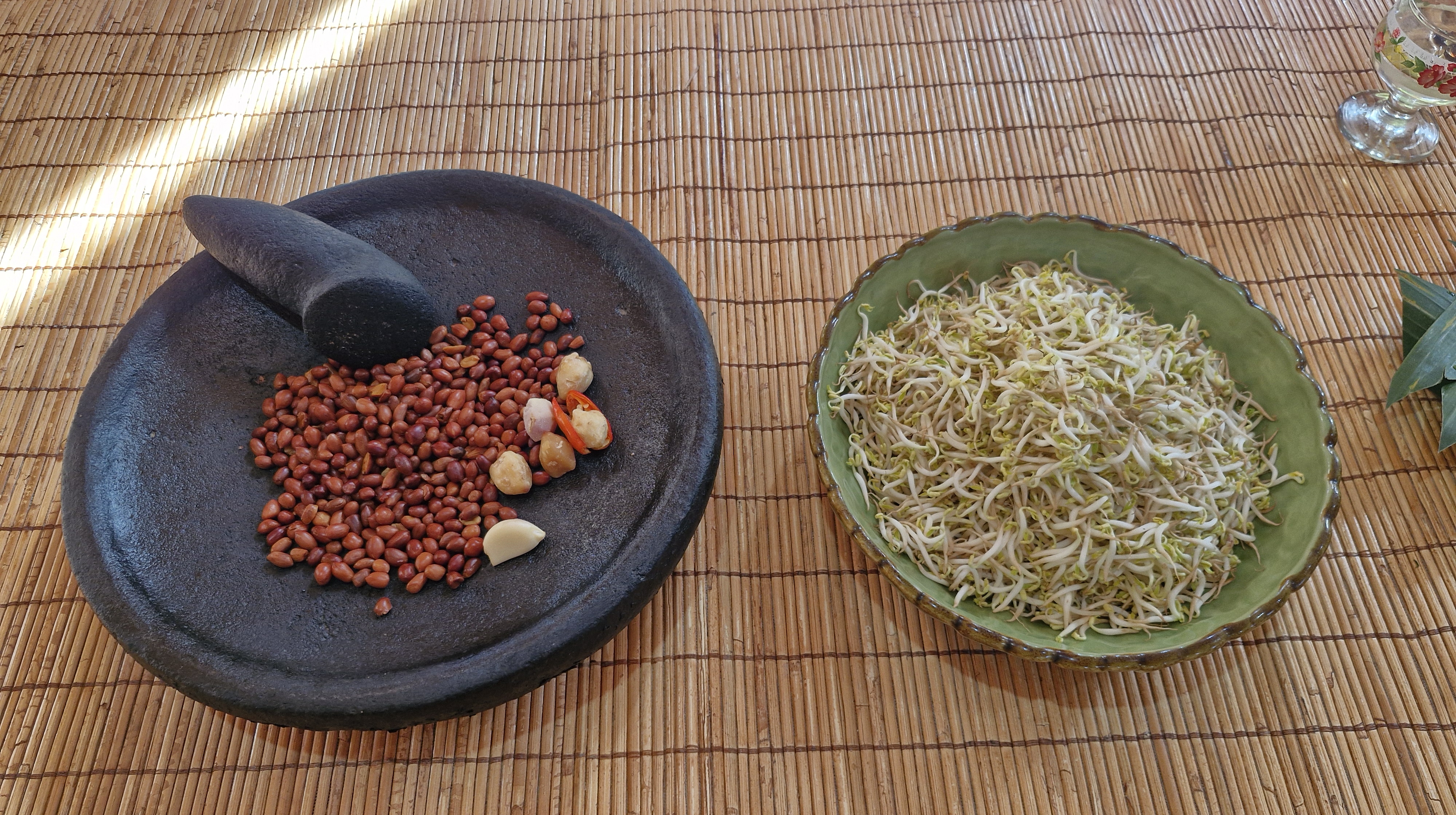 A stone mortar and pestle with red beans, garlic, and chili peppers beside a bowl of bean sprouts on a bamboo mat.