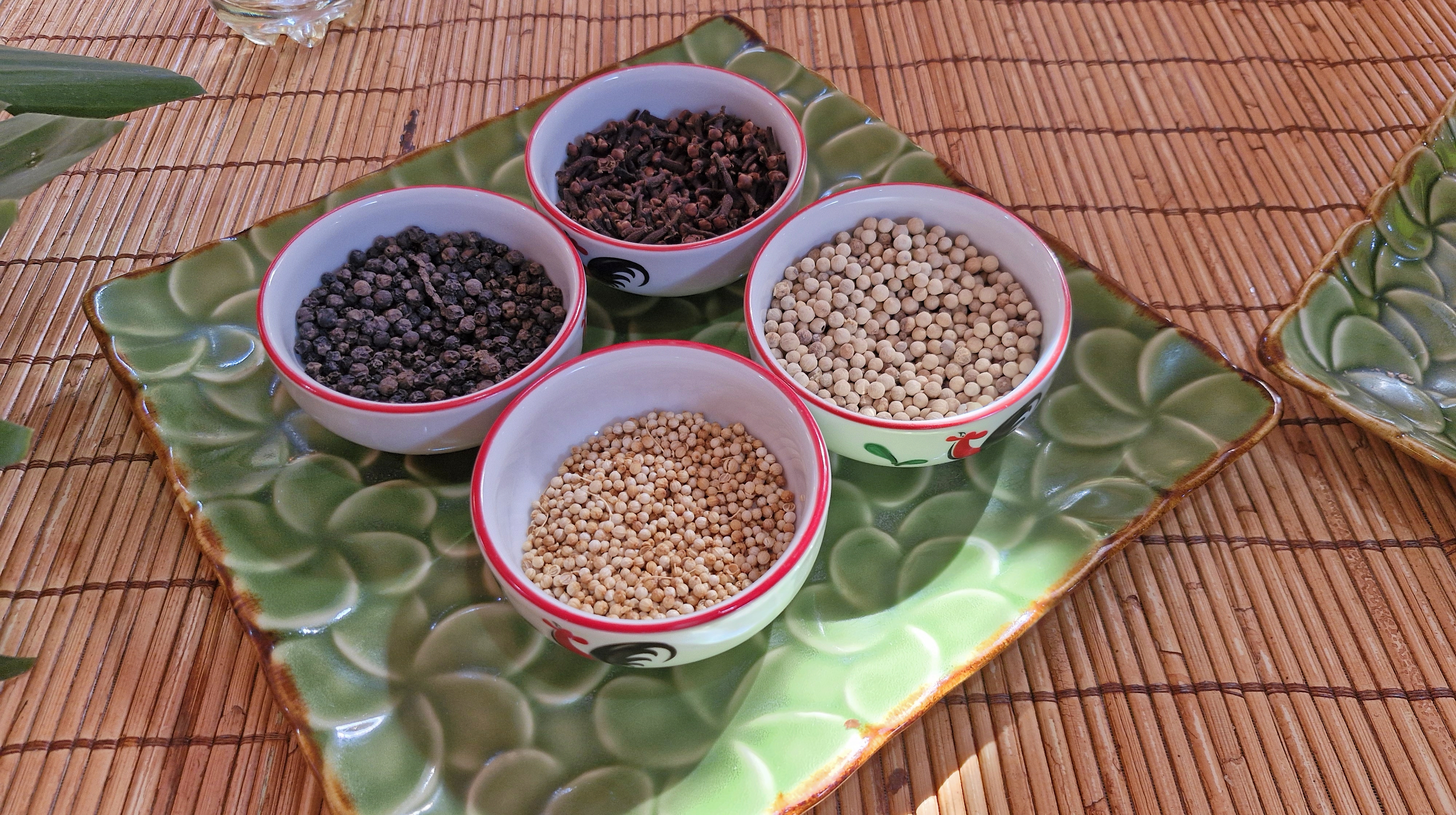 A selection of spices in small bowls on a decorative green plate, placed on a bamboo mat. The bowls contain black peppercorns, white pepper, mustard seeds, and dried cloves.