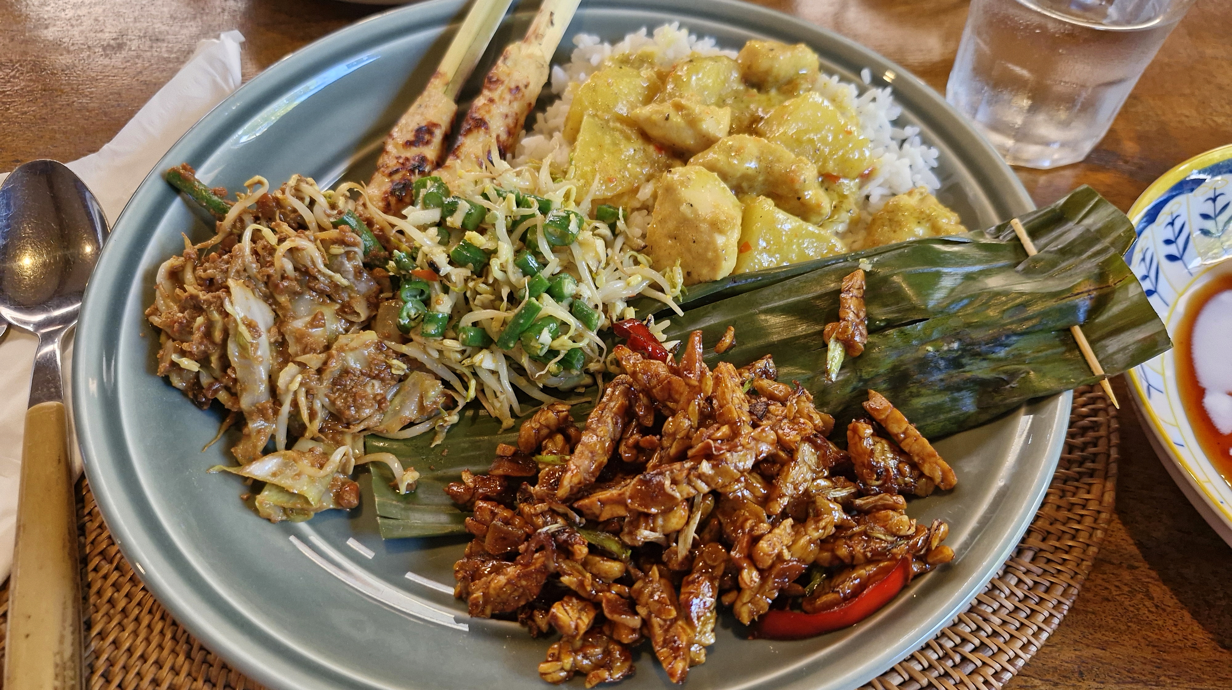 A plate of Indonesian cuisine featuring rice topped with a yellow curry, various vegetables, chicken skewers, and a mix of side dishes including shredded cabbage and fried tempeh.