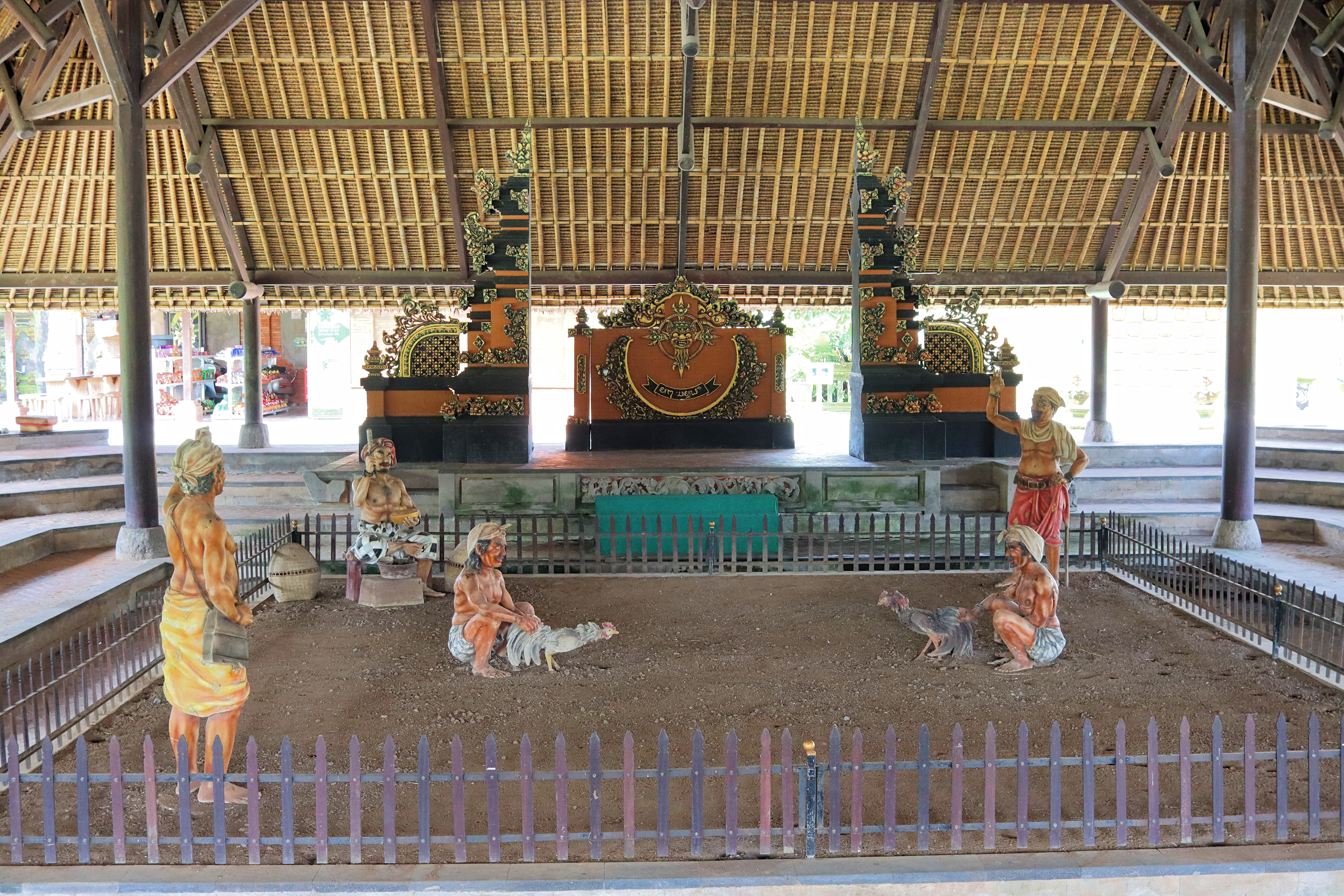 An indoor setting featuring a traditional Balinese structure with a thatched roof, surrounded by ornate decorations. In the center, there are life-sized statues depicting men involved in a cockfighting scene, with a fenced area and seating around the perimeter.