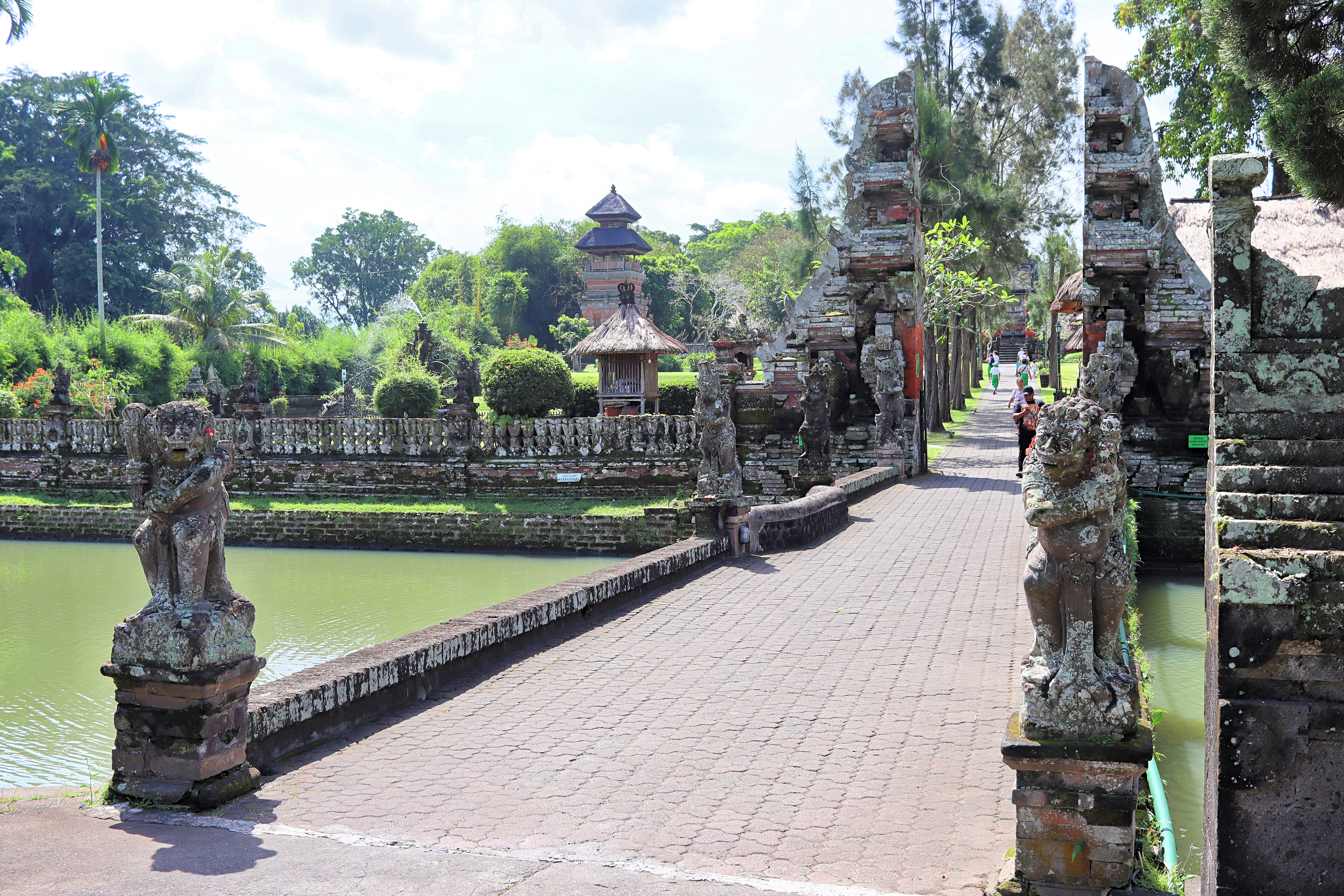 A serene view of a Balinese temple complex featuring a stone bridge leading across a pond, with intricately carved statues and lush greenery in the background.