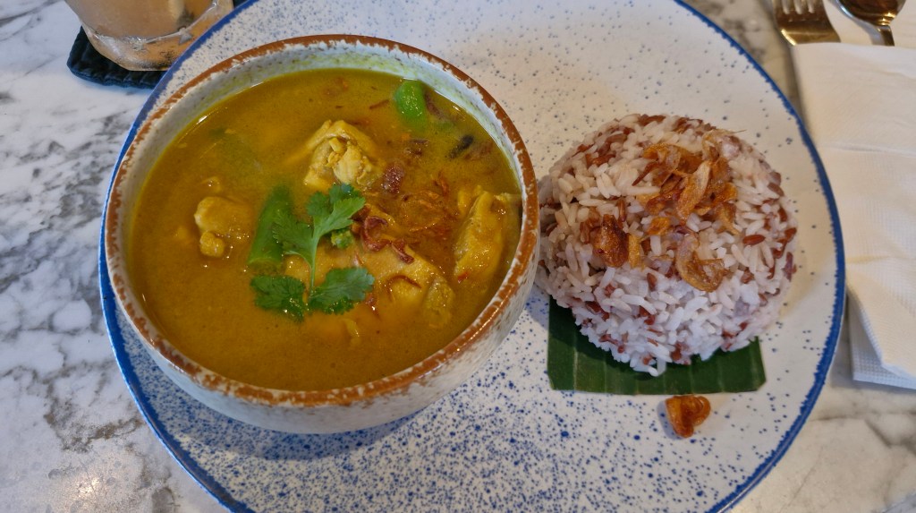 A colorful bowl of chicken curry served with red rice, garnished with cilantro and fried shallots, on a marble table.