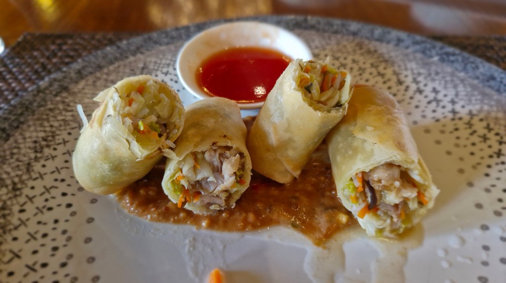 A plate of spring rolls filled with vegetables and meat, accompanied by a small bowl of red dipping sauce.