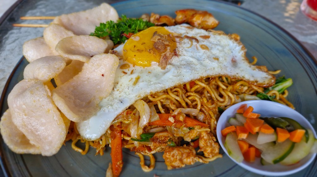 A plate of fried noodles topped with a sunny-side-up egg, accompanied by crispy crackers and a small bowl of pickled vegetables.