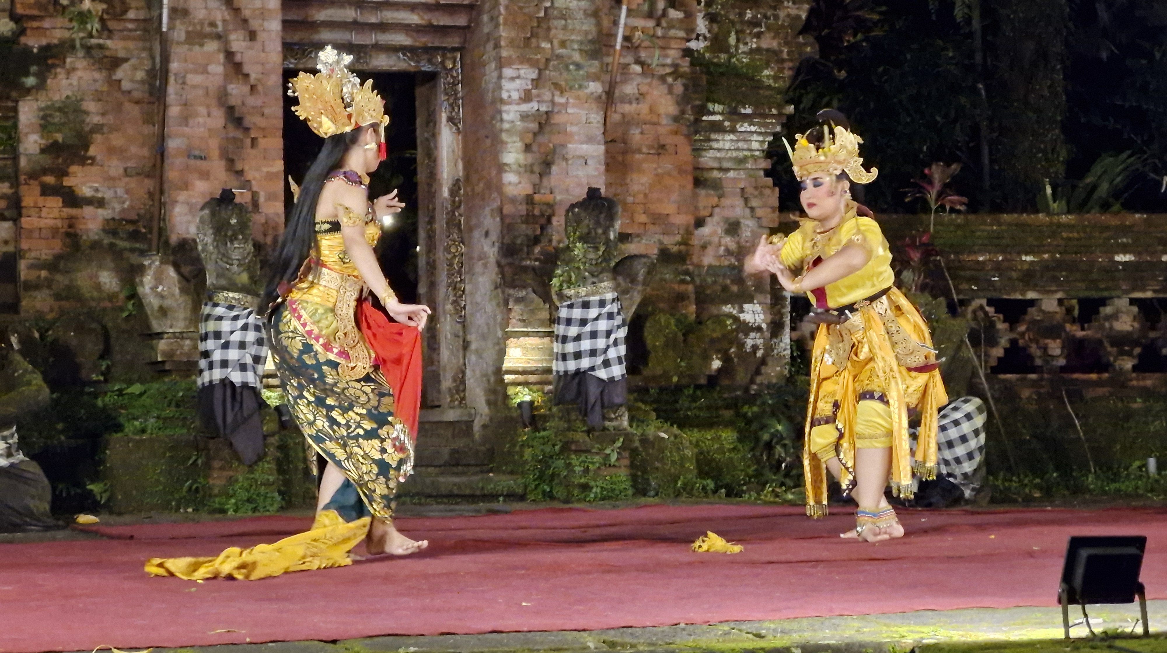 Two performers in traditional Balinese costumes are dancing on stage during the Ramayana Ballet at the ARMA Museum. The background features stone structures and lush greenery.