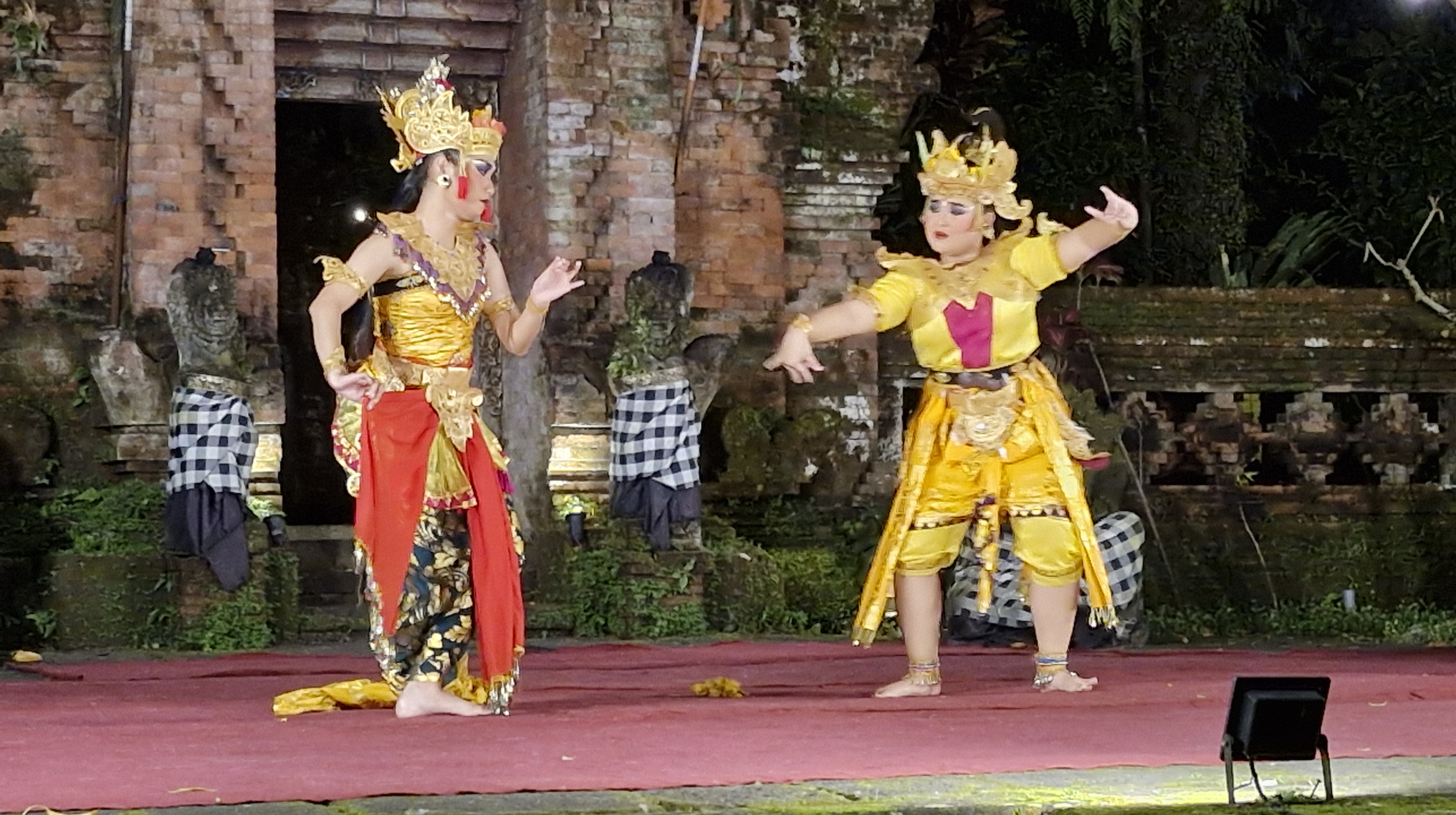Two performers in traditional Balinese costumes dancing during a Ramayana Ballet performance, with a historical temple backdrop.