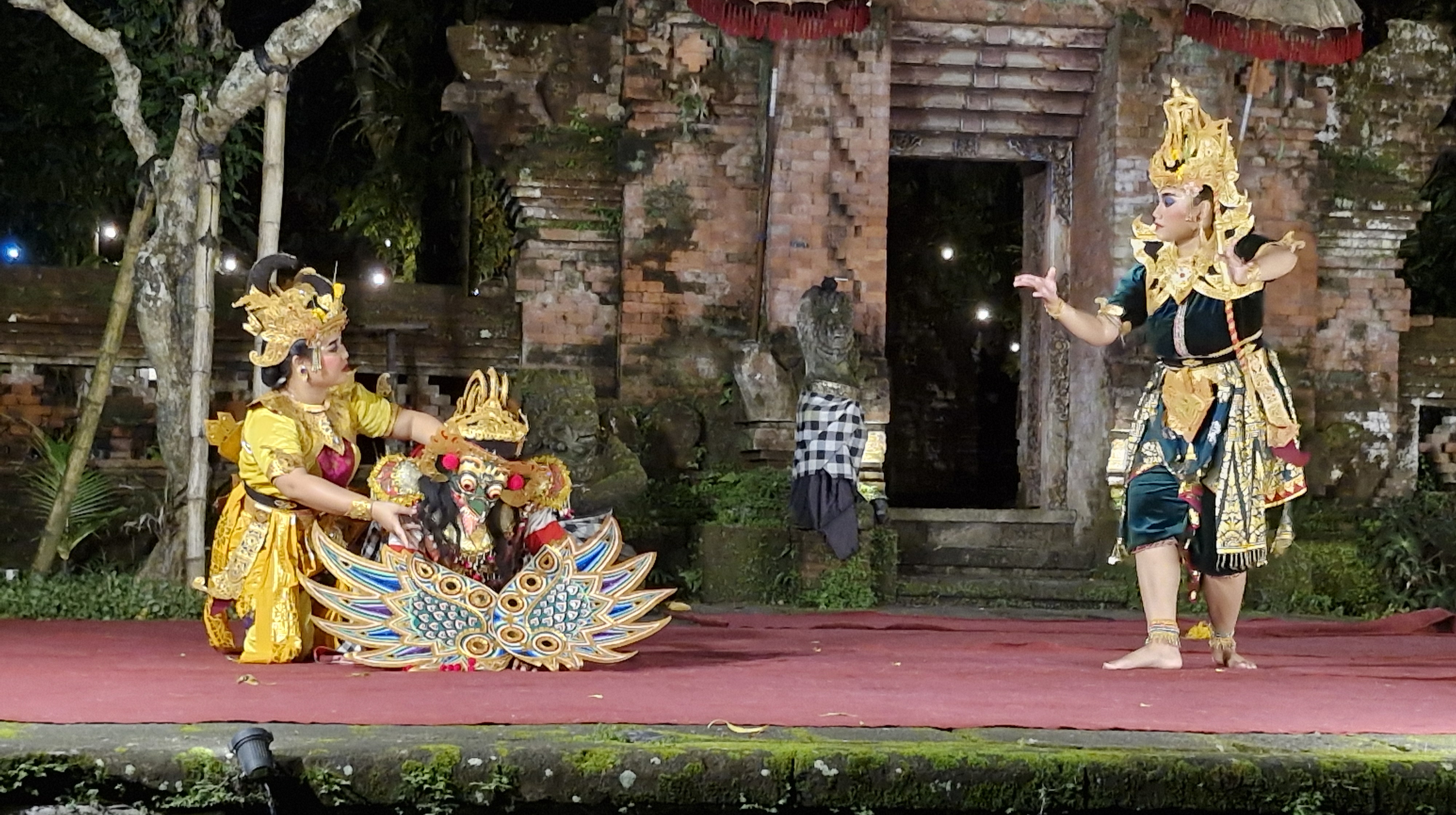 Two performers dressed in traditional Balinese costume are acting on stage during a cultural dance performance. One performer holds a colorful mask while the other gestures dramatically, set against a backdrop of ancient temple ruins illuminated by soft lighting.