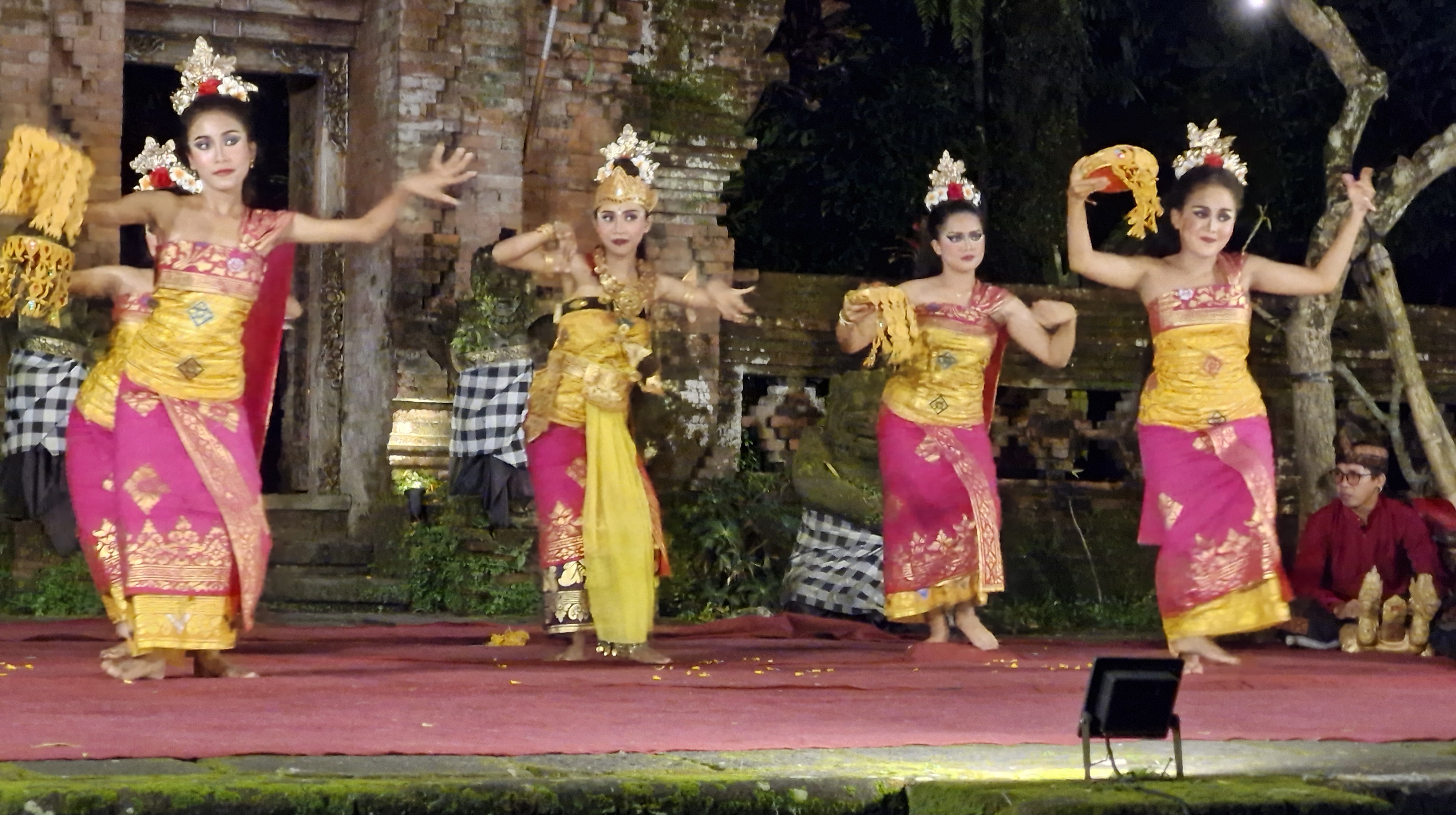 Four female dancers performing the Ramayana Ballet in traditional Balinese costumes with intricate headdresses, set against a temple backdrop.