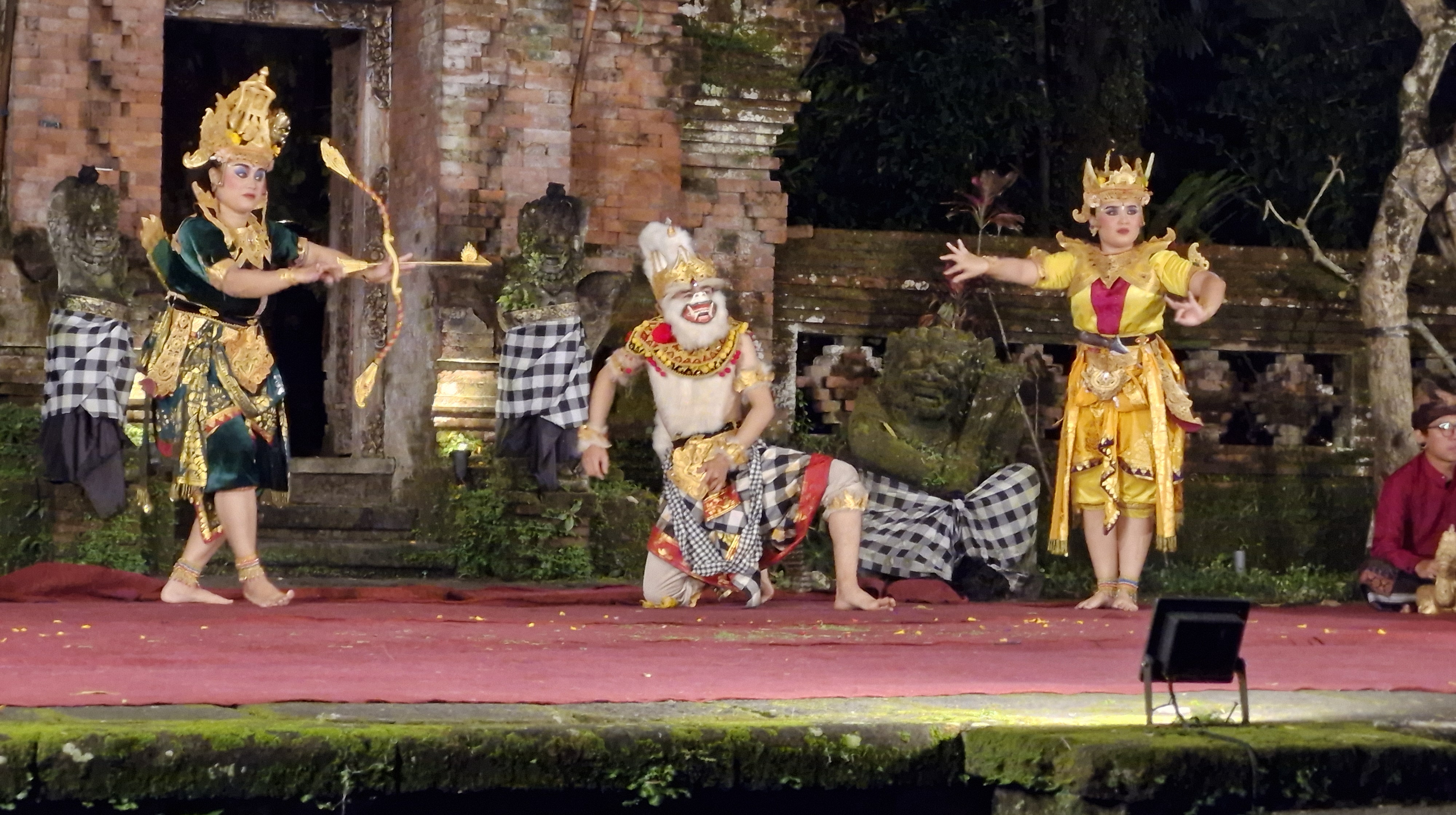 A scene from the Ramayana Ballet performance, showcasing three dancers in traditional Balinese costumes on stage, with a backdrop of ancient temple ruins.