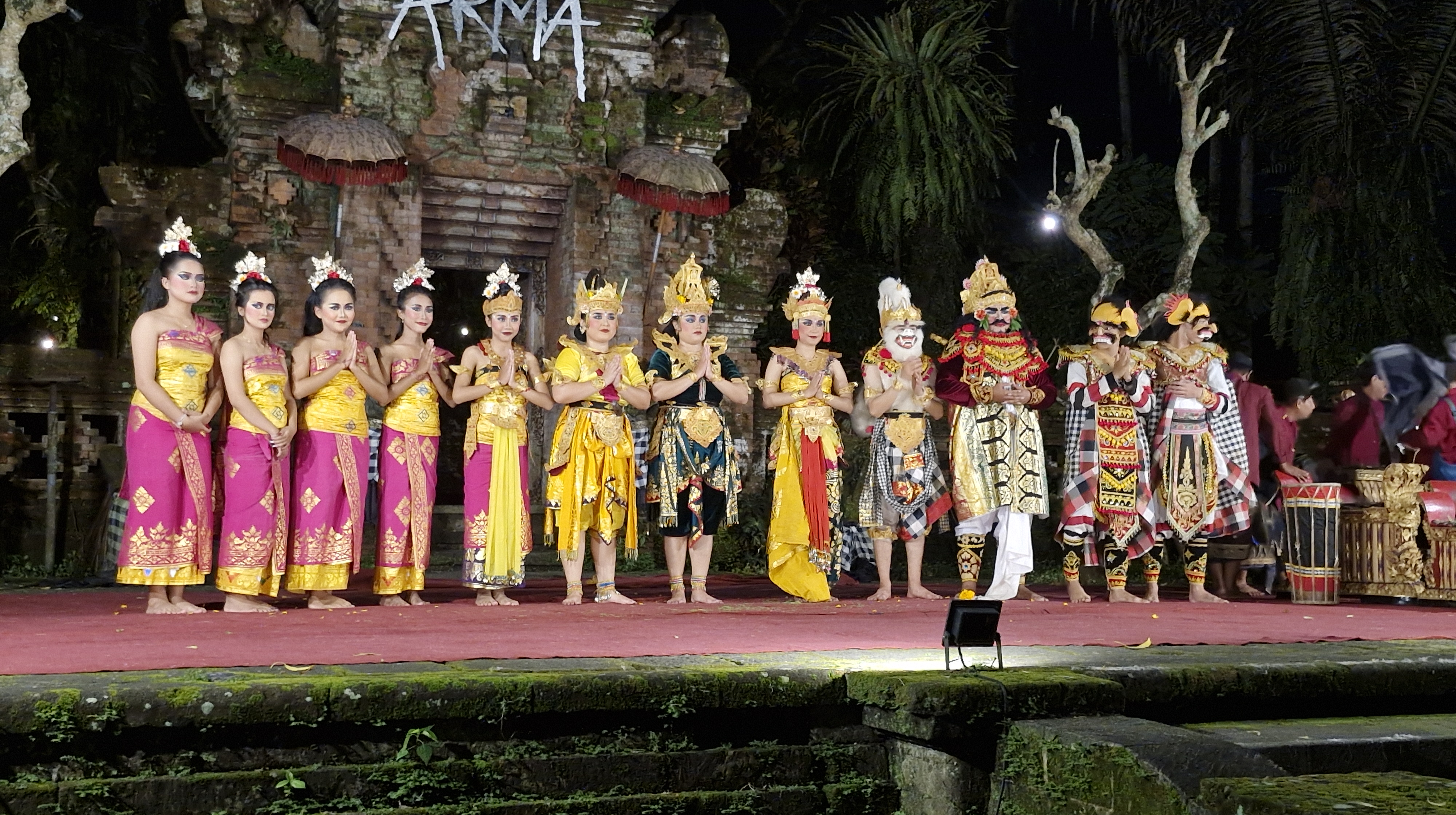 A group of performers in traditional Balinese costumes stand on stage at night, preparing for the Ramayana Ballet at the ARMA Museum.
