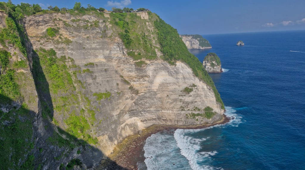 A breathtaking view of a steep, lush cliffside overlooking the vibrant blue ocean. Waves crash against the rocky shore below, creating a picturesque coastal scene.