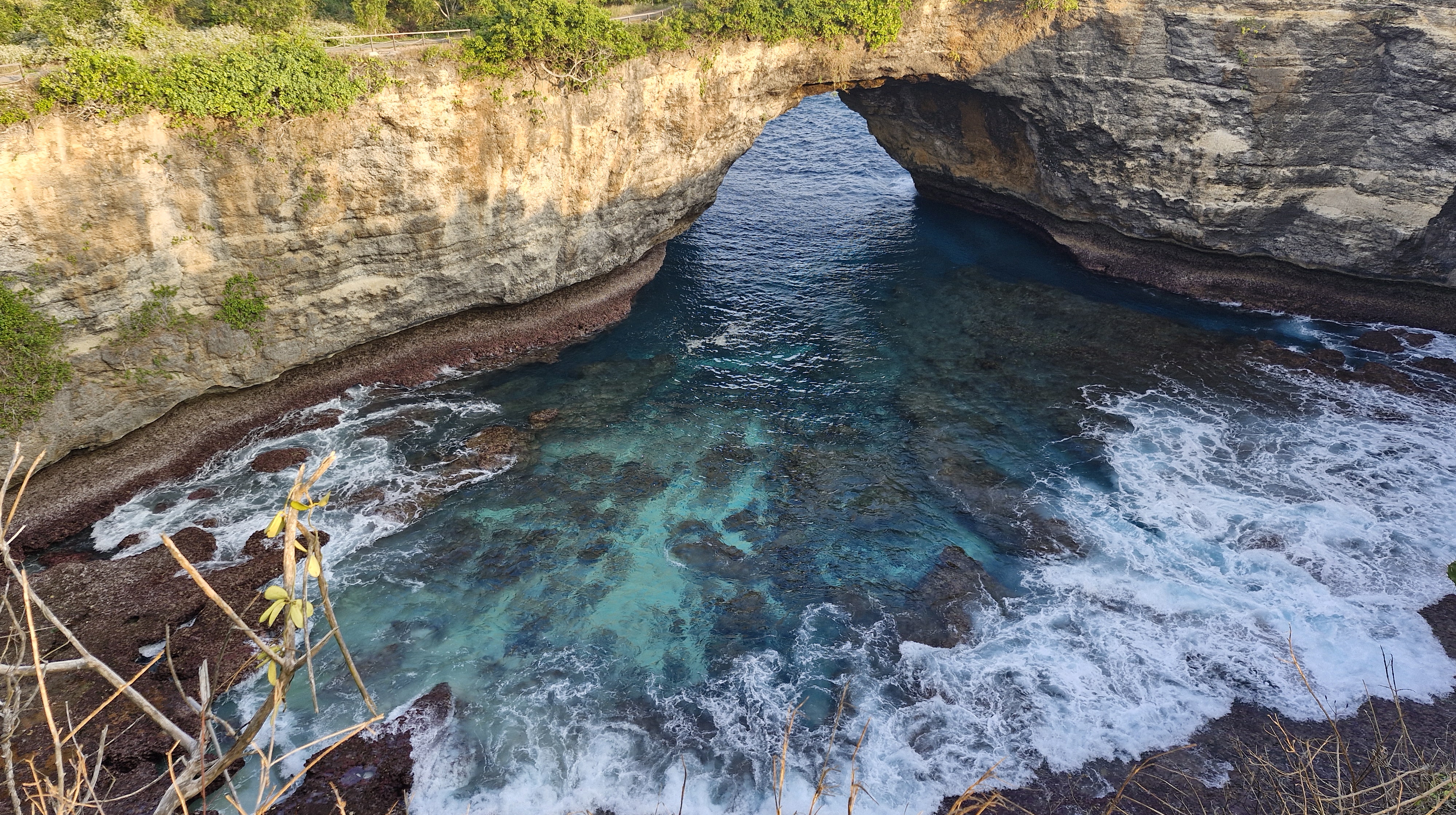 A view of Broken Beach on Nusa Penida, featuring a natural arch in the rocky cliffs with turquoise waters below and white waves crashing against the shoreline.