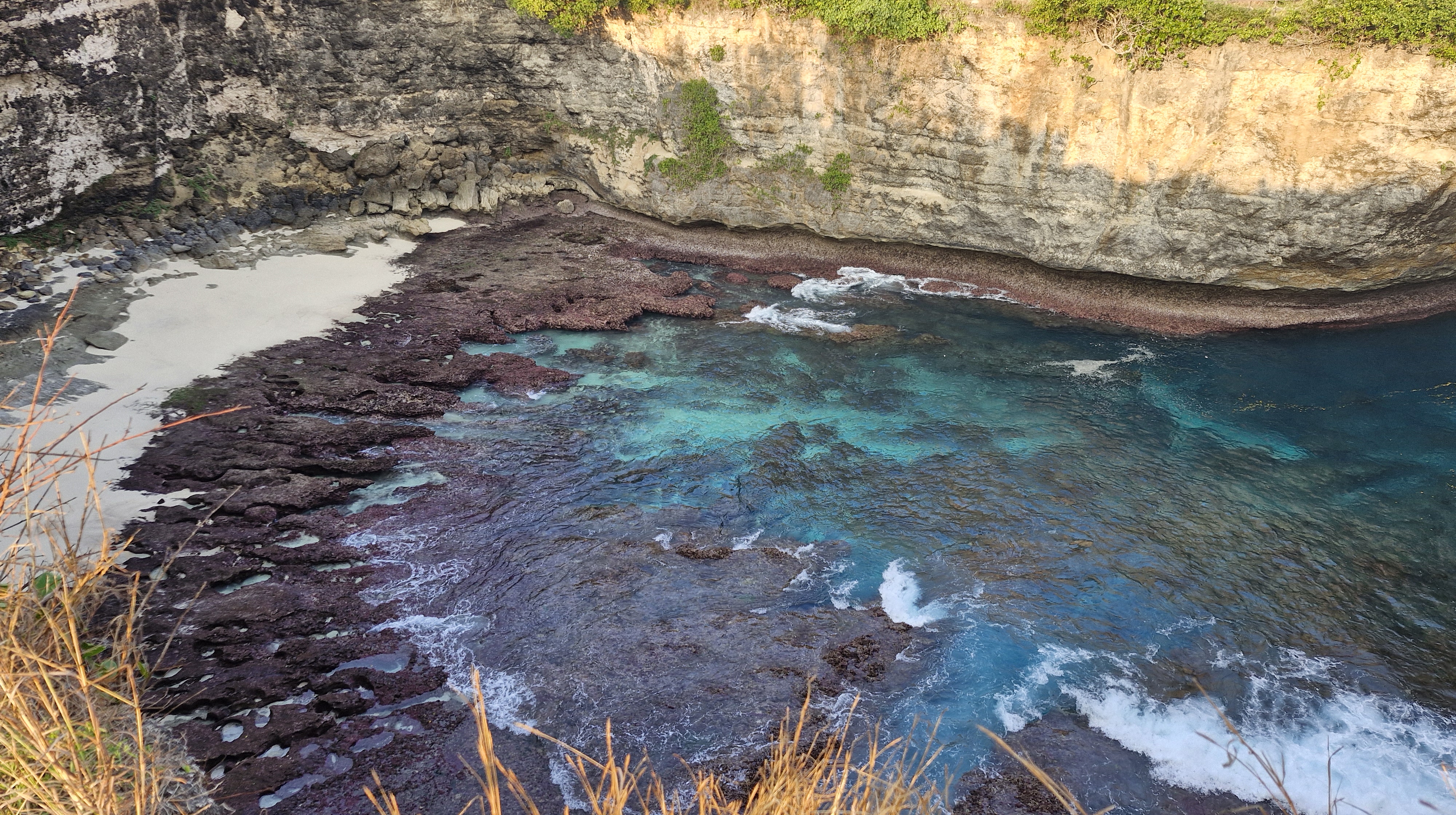 Aerial view of a rocky cove with turquoise waters and a sandy beach section, surrounded by steep cliffs covered in greenery.