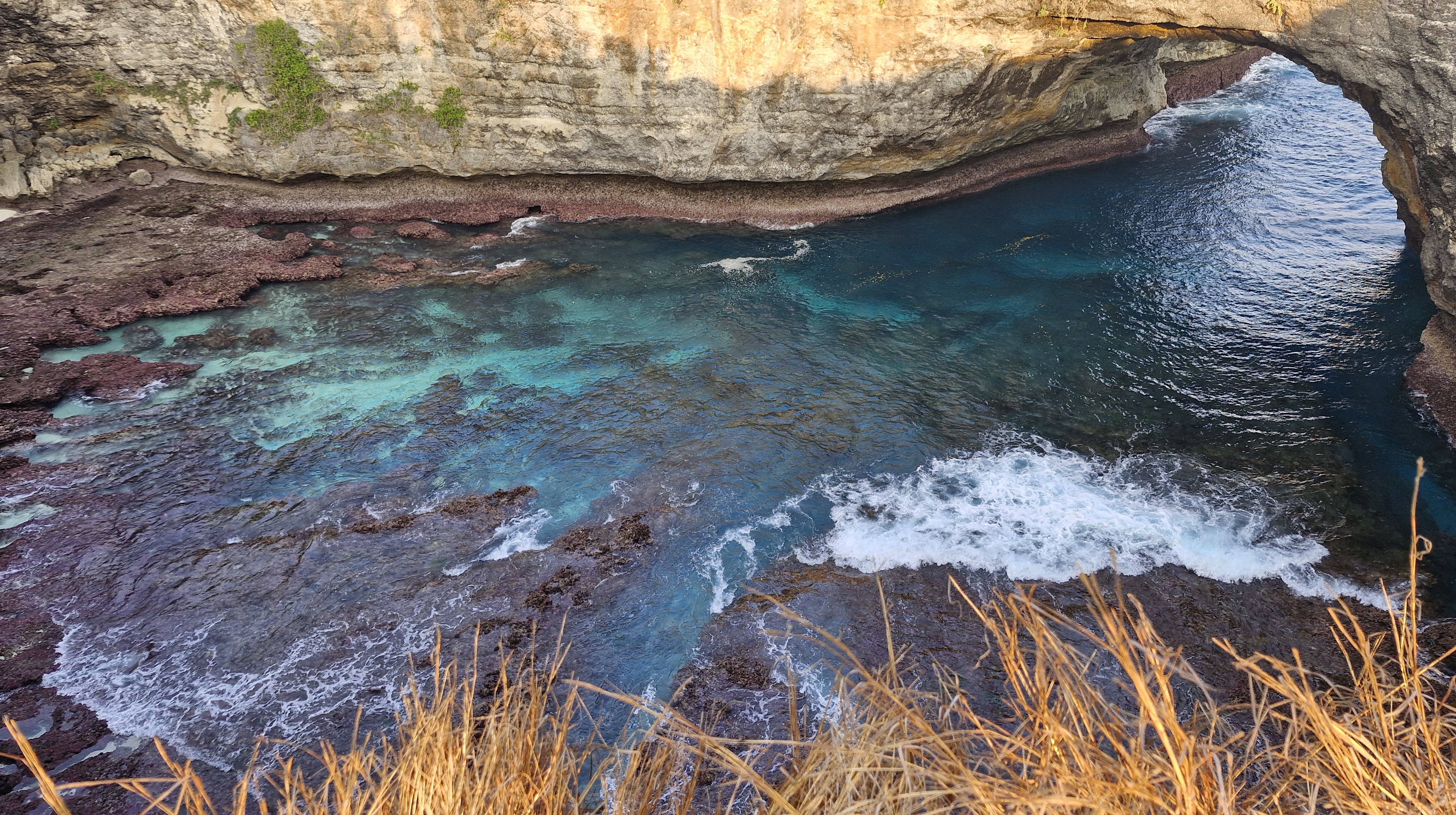 Aerial view of crystal-clear turquoise water crashing against rocky shores near a cliff arch.