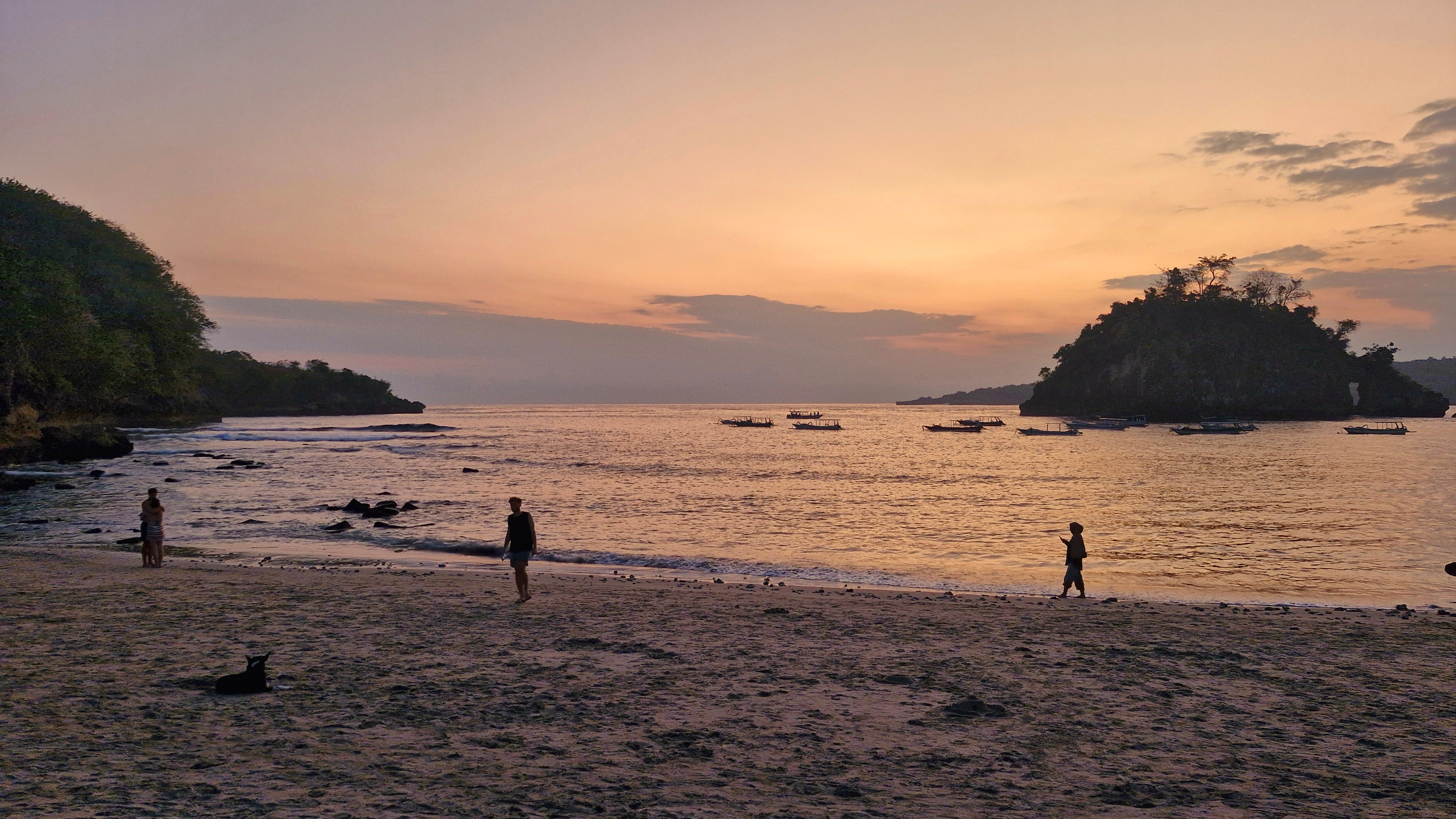A sunset view at a beach with silhouettes of people walking on the sand and boats in the calm water.