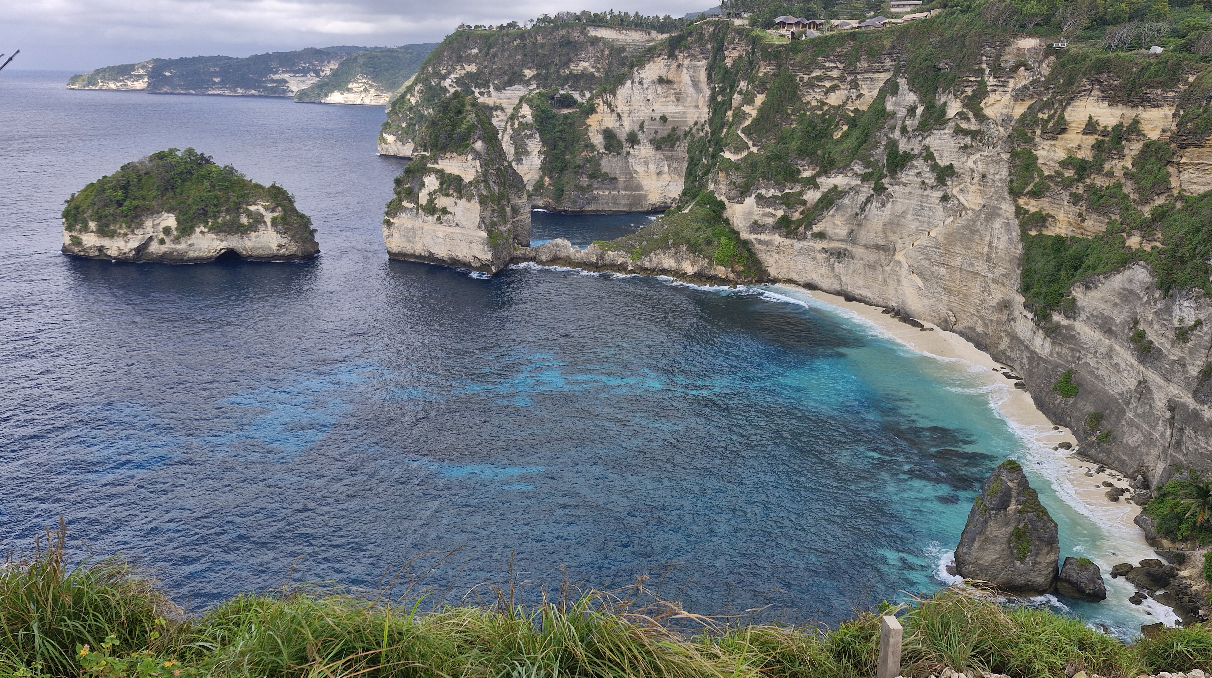 A panoramic view of Kelingking Beach in Nusa Penida, showcasing dramatic cliffs, turquoise waters, and a small rocky island in the foreground.