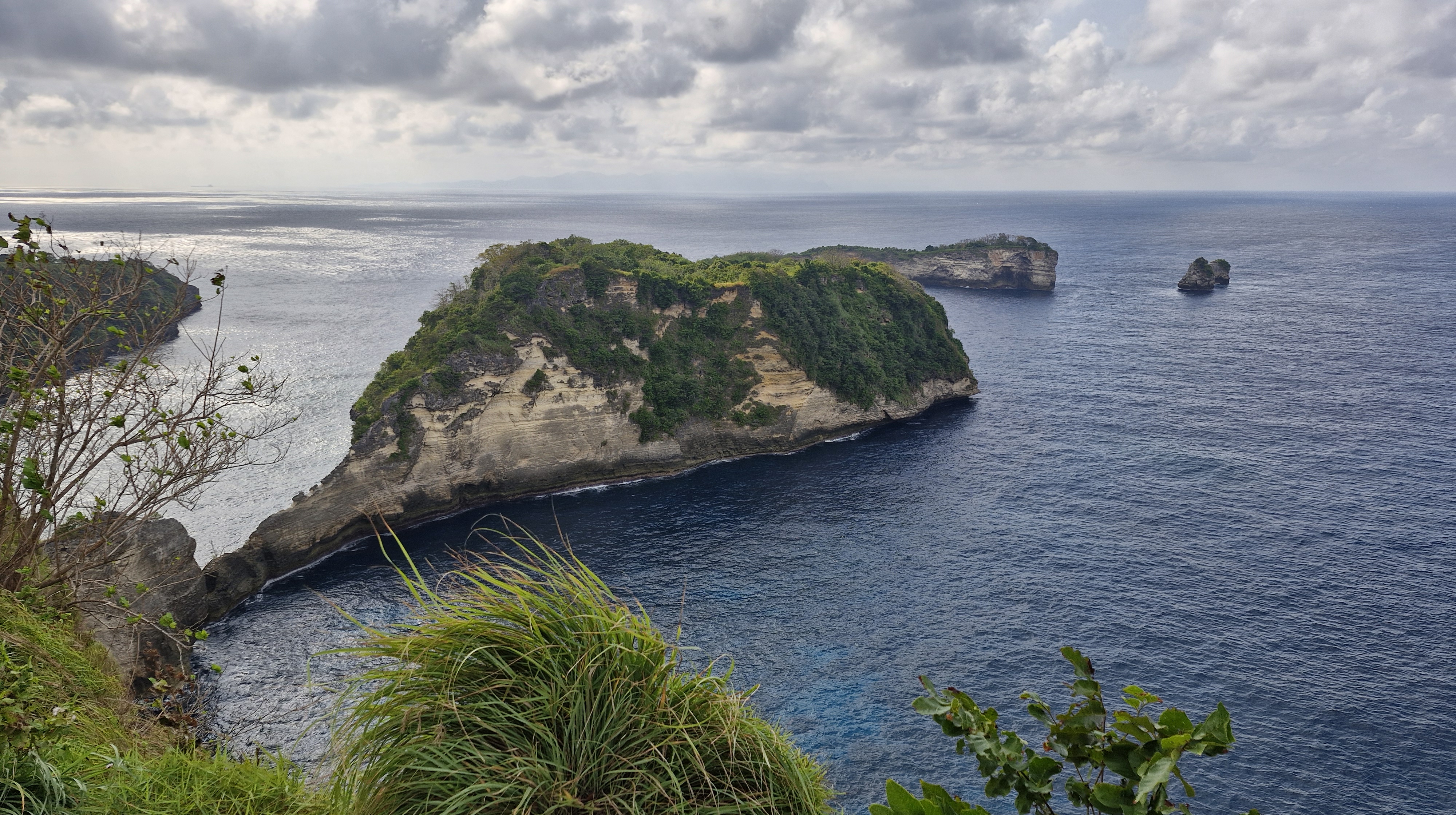 A scenic view of cliffs and rocky outcrops along the coastline of Nusa Penida, with lush greenery and a calm ocean under a cloudy sky.