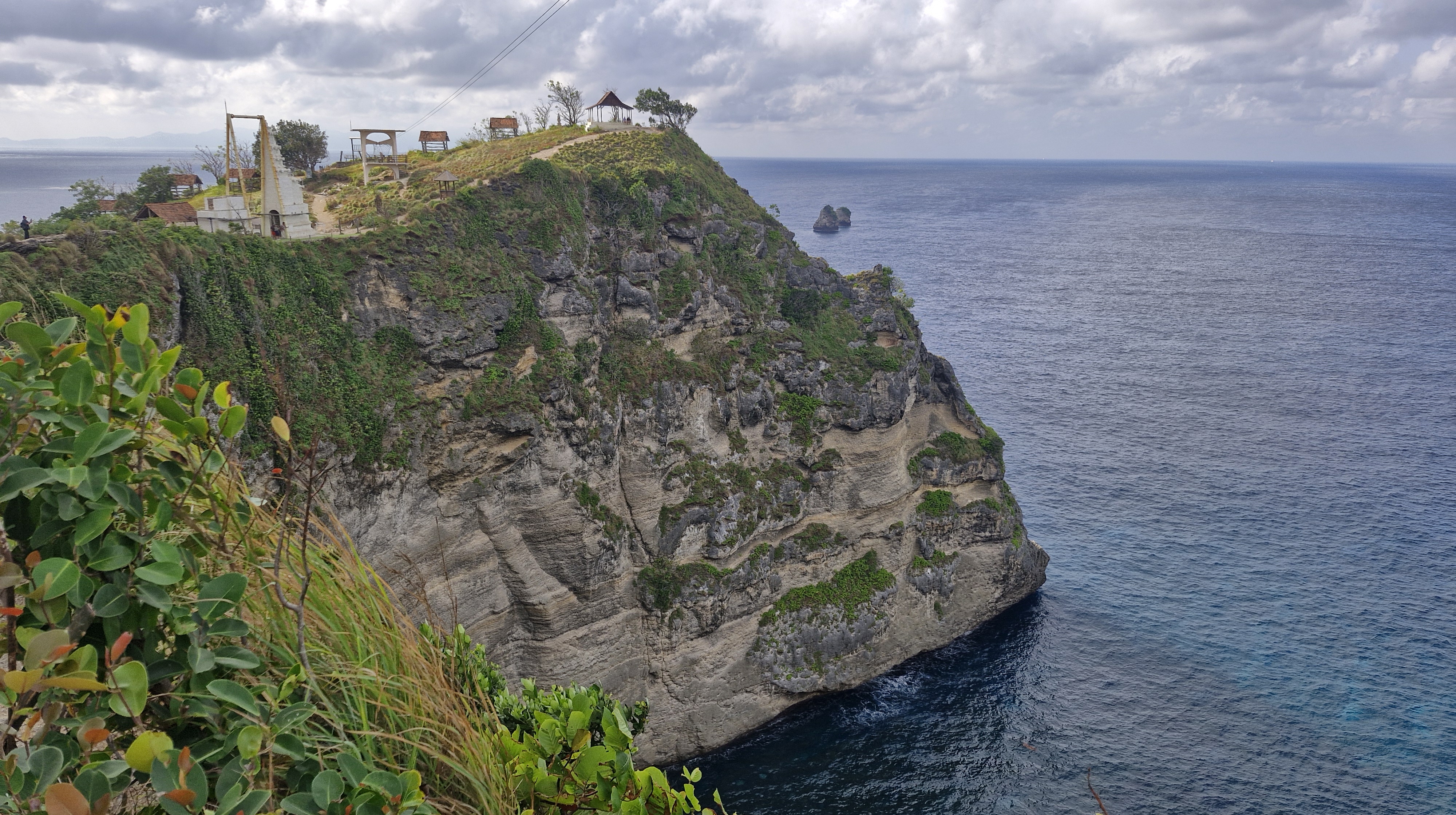 A panoramic view of a cliff landscape overlooking a calm ocean, featuring lush green vegetation along the rocky cliffside and a gathering area with small structures on top.