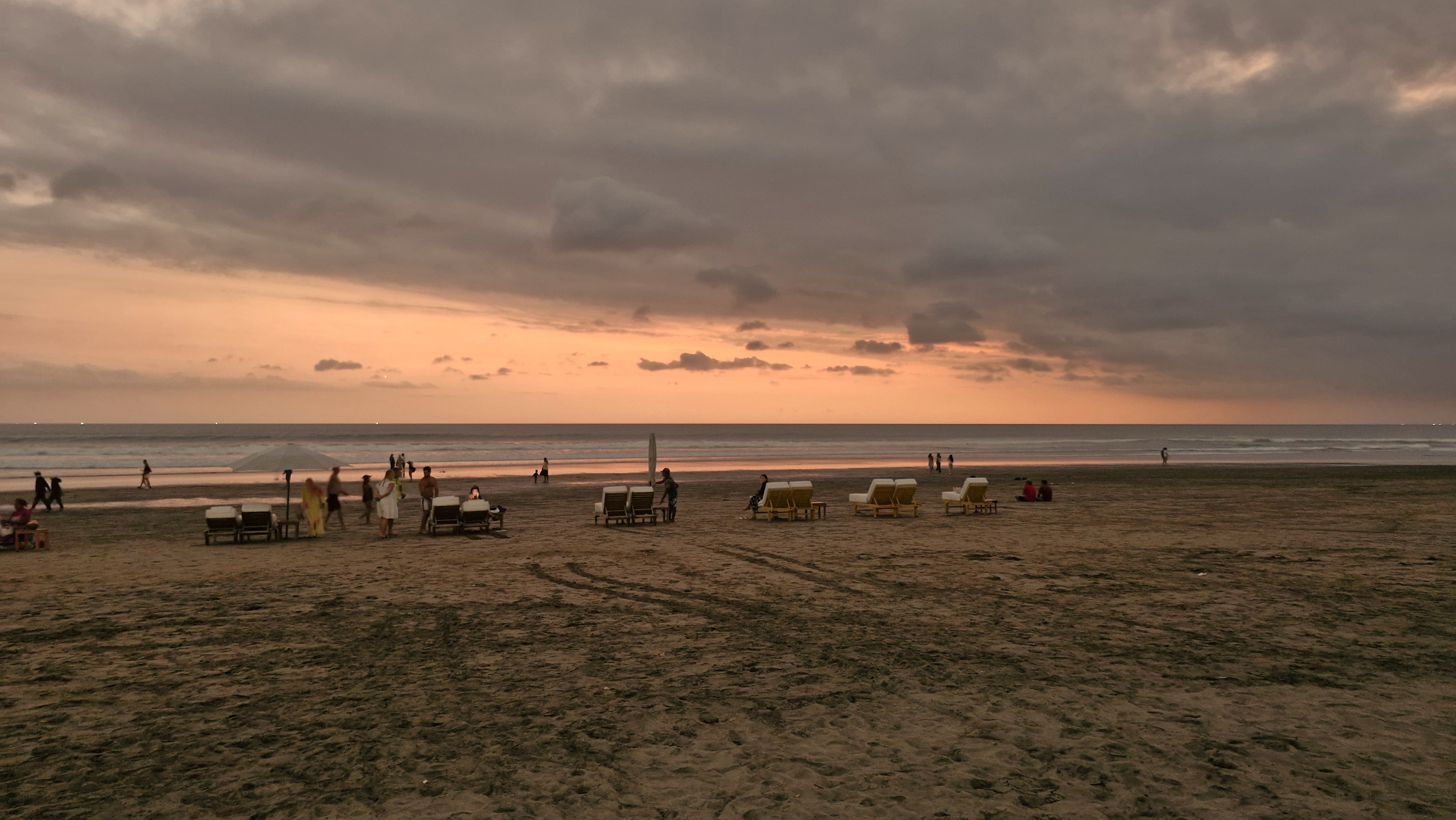 A beach scene at sunset with people walking along the shore. Empty lounge chairs are scattered on the sand, and the sky features a blend of warm colors.