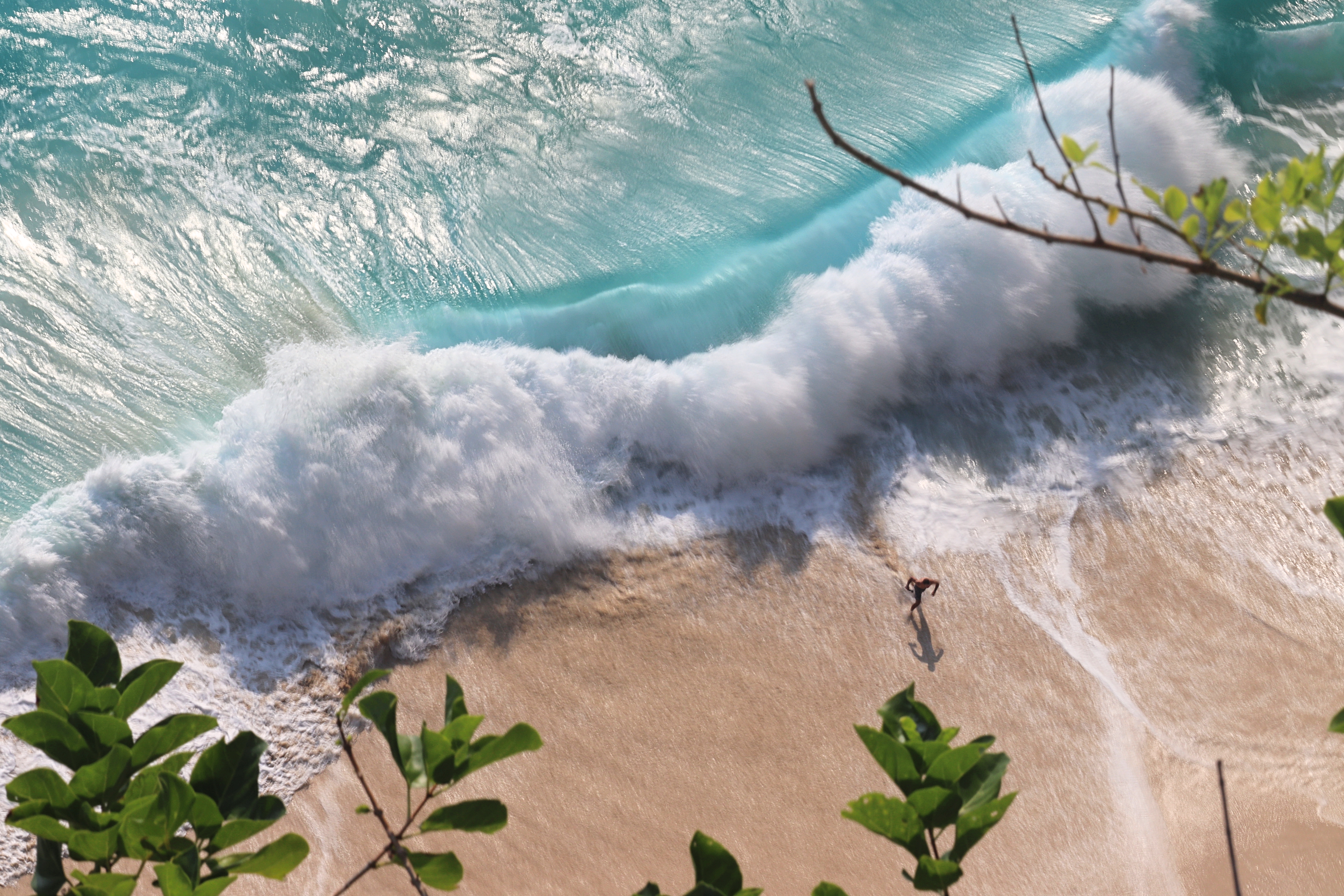 Aerial view of a person walking on a sandy beach with waves crashing nearby, surrounded by clear turquoise water.